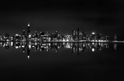 A distant band of lit towers sits low in the frame and repeats almost exactly in the flat water beneath it, sky and shore both pure black.