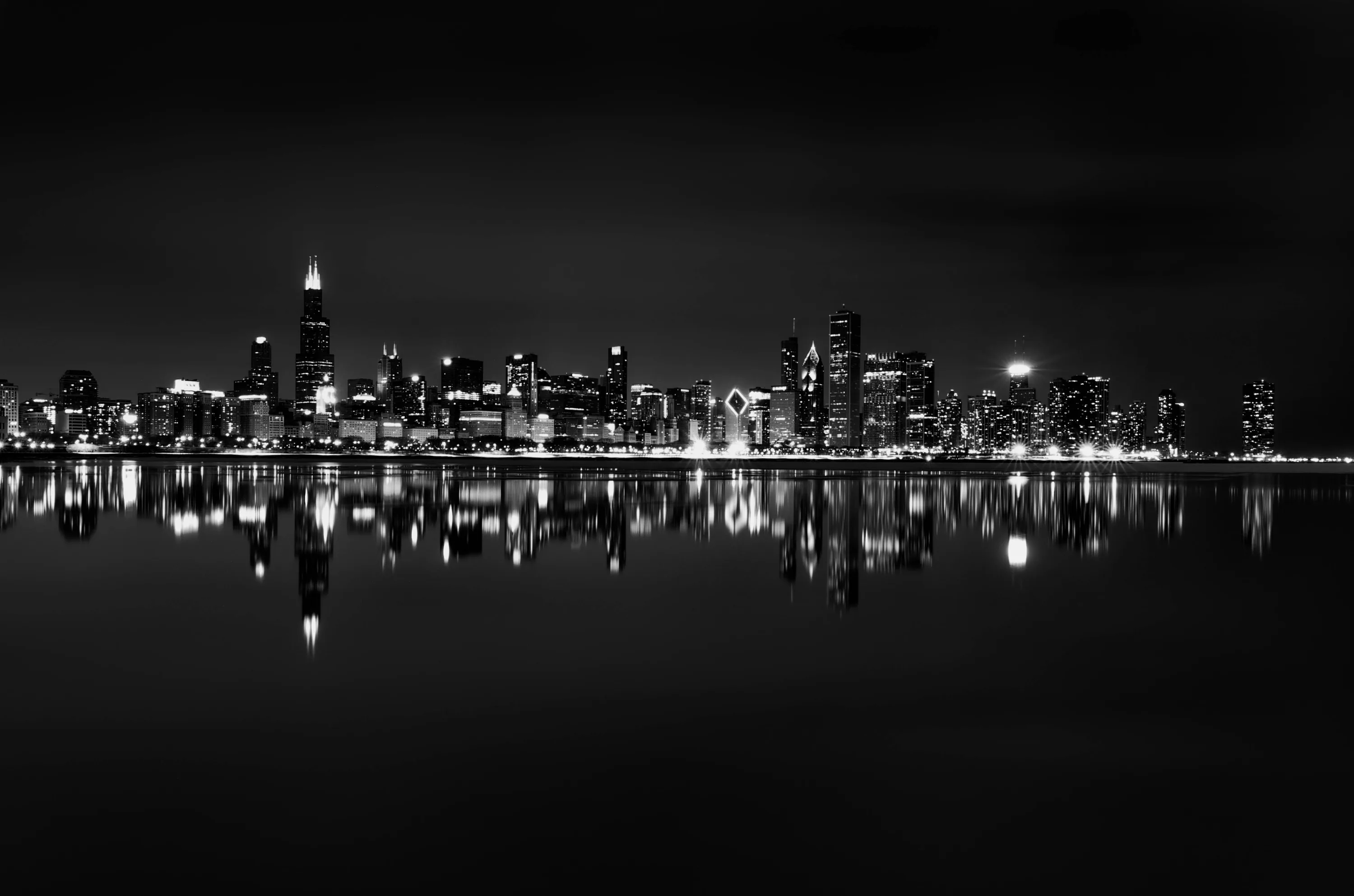 A distant band of lit towers sits low in the frame and repeats almost exactly in the flat water beneath it, sky and shore both pure black.