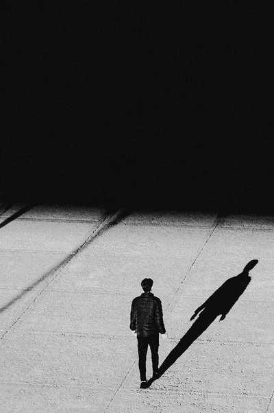 One man crosses pale paving while his stretched shadow doubles him to the right, the whole upper half solid black where a clock can sit.
