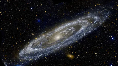 The Andromeda galaxy tilts across a field of foreground stars, a yellow core wrapped in blue arms with a small companion galaxy below.