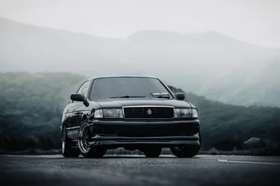 A boxy black saloon on deep-dish wheels sits head-on, headlights off, layered ridges and low cloud filling the whole background.