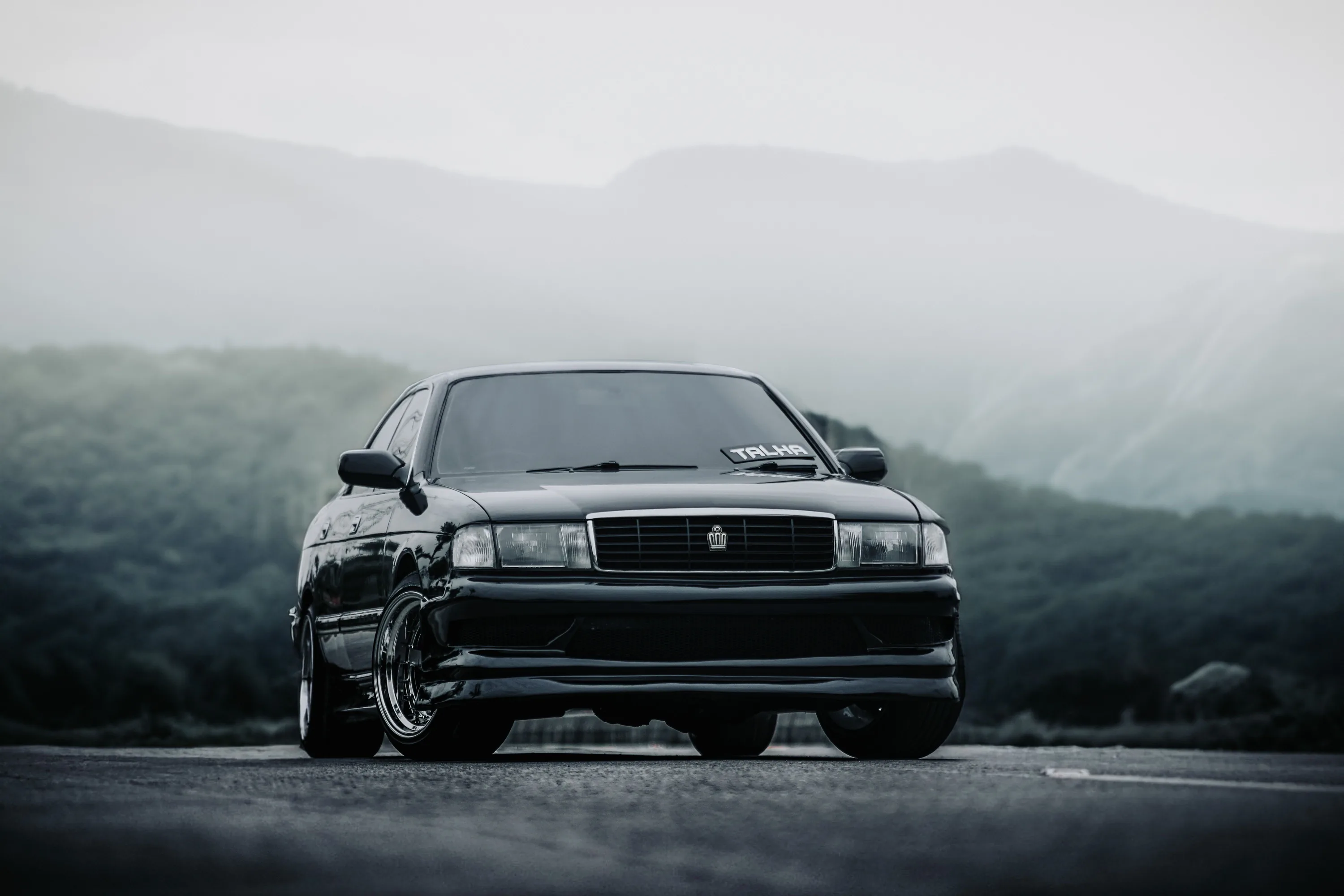 A boxy black saloon on deep-dish wheels sits head-on, headlights off, layered ridges and low cloud filling the whole background.