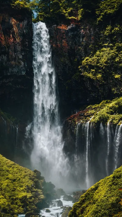 Spray drifts off a high single-drop waterfall flanked by smaller cascades, moss covering the rock on either side; the tall frame suits a phone without cropping.