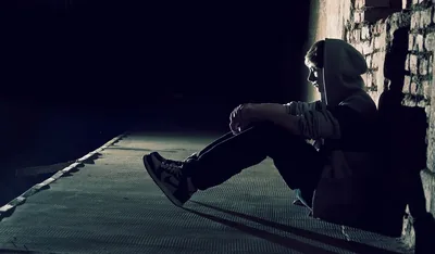 A person in a hoodie sits on a metal walkway leaning on brick, lit hard from the left so long shadows rake across the grating.