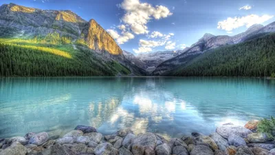 Turquoise glacial water stretches to the Victoria Glacier under scattered cloud, rounded stones lining the near shore; the smooth mid-lake area takes a clock well.