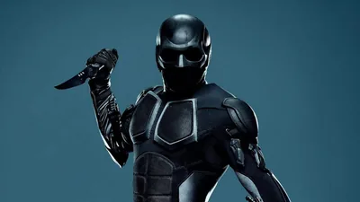 A full-body shot of a black armoured suit and helmet, knife reversed in one hand, set against a flat teal-grey backdrop.