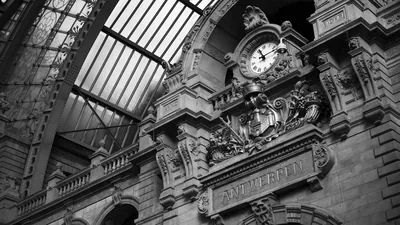 An ornate railway clock sits above carved stonework and an iron roof, shot in high-contrast monochrome.