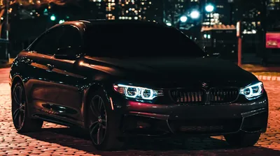 Front three-quarter on cobbles, corona headlights lit and a long string of out-of-focus office windows and streetlights stretching away behind the roofline.