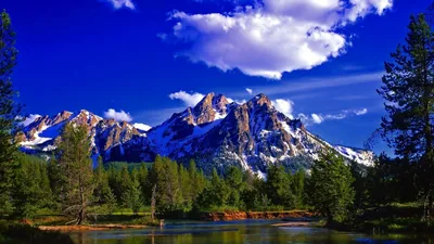 Saturated blue sky and bright cumulus tower over rugged peaks, a quiet river and pine forest filling the foreground.