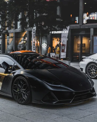 A matte black Lamborghini parked kerbside on paving stones, shop windows and a white sports car reflected in its glossy quarter panel and forged wheels.