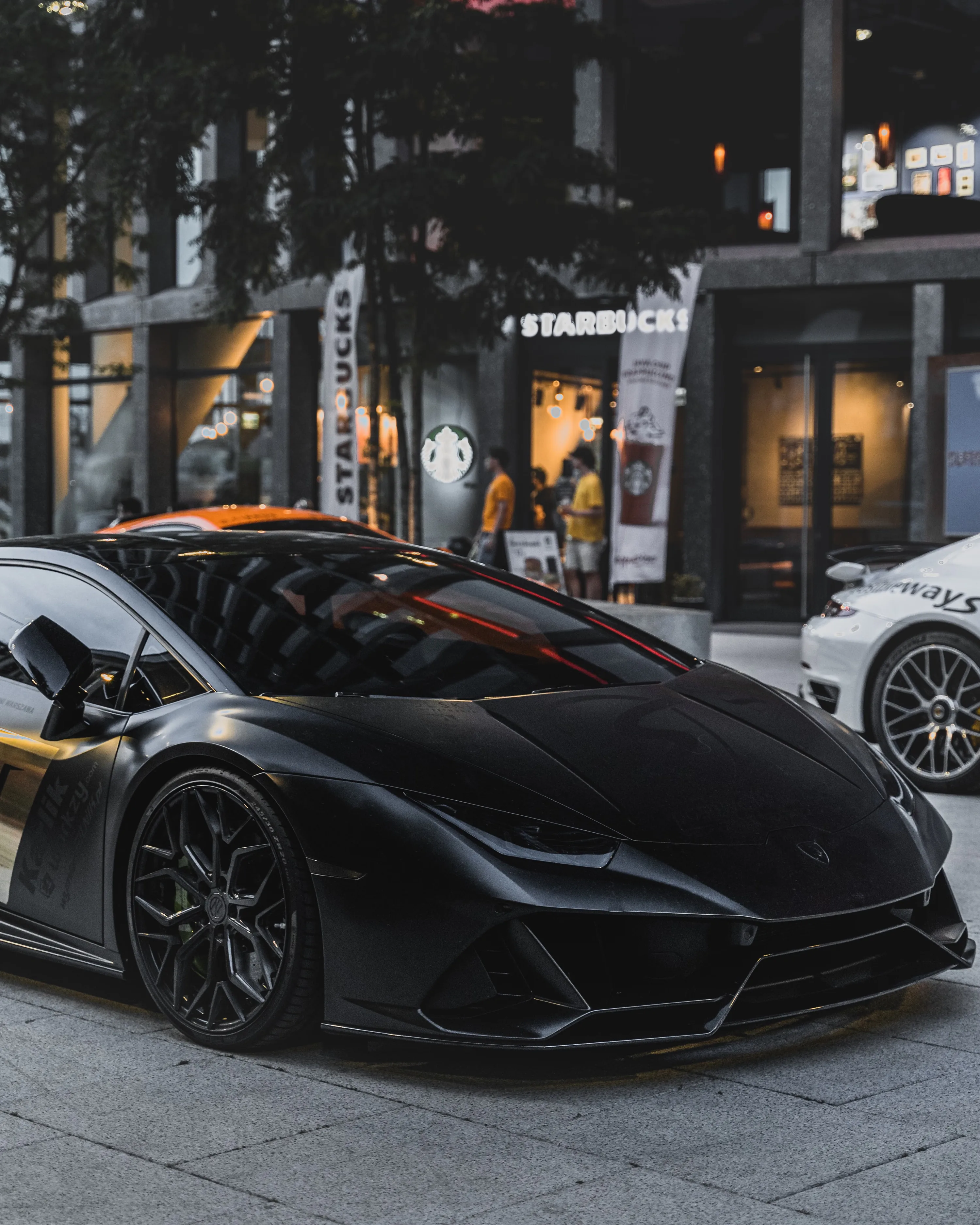 A matte black Lamborghini parked kerbside on paving stones, shop windows and a white sports car reflected in its glossy quarter panel and forged wheels.