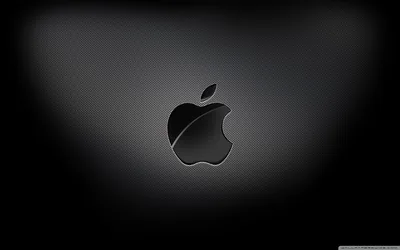 The Apple mark sits glossy at the centre of a fine carbon weave, a soft spotlight behind it falling away to black corners.