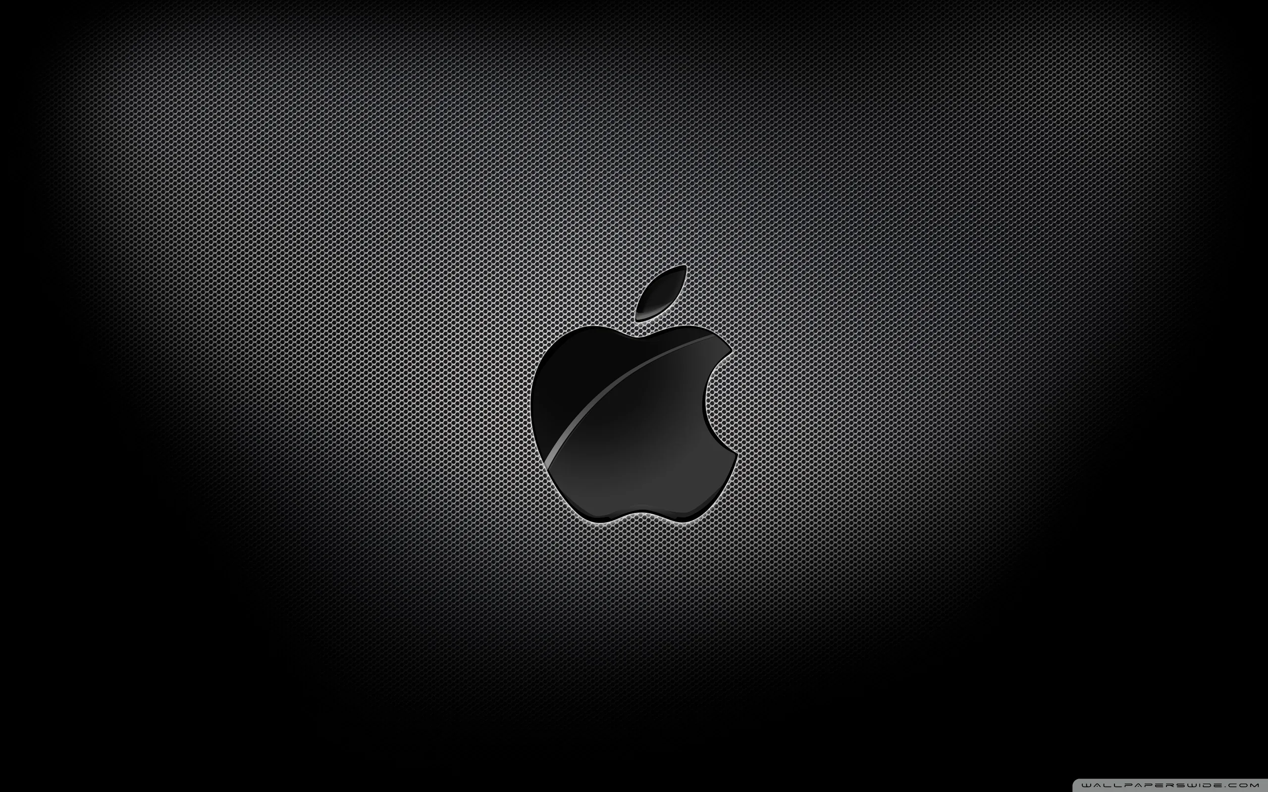 The Apple mark sits glossy at the centre of a fine carbon weave, a soft spotlight behind it falling away to black corners.