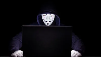 A hooded figure in a pale grinning mask sits behind an open laptop, hands on the desk and the room around them fully black.