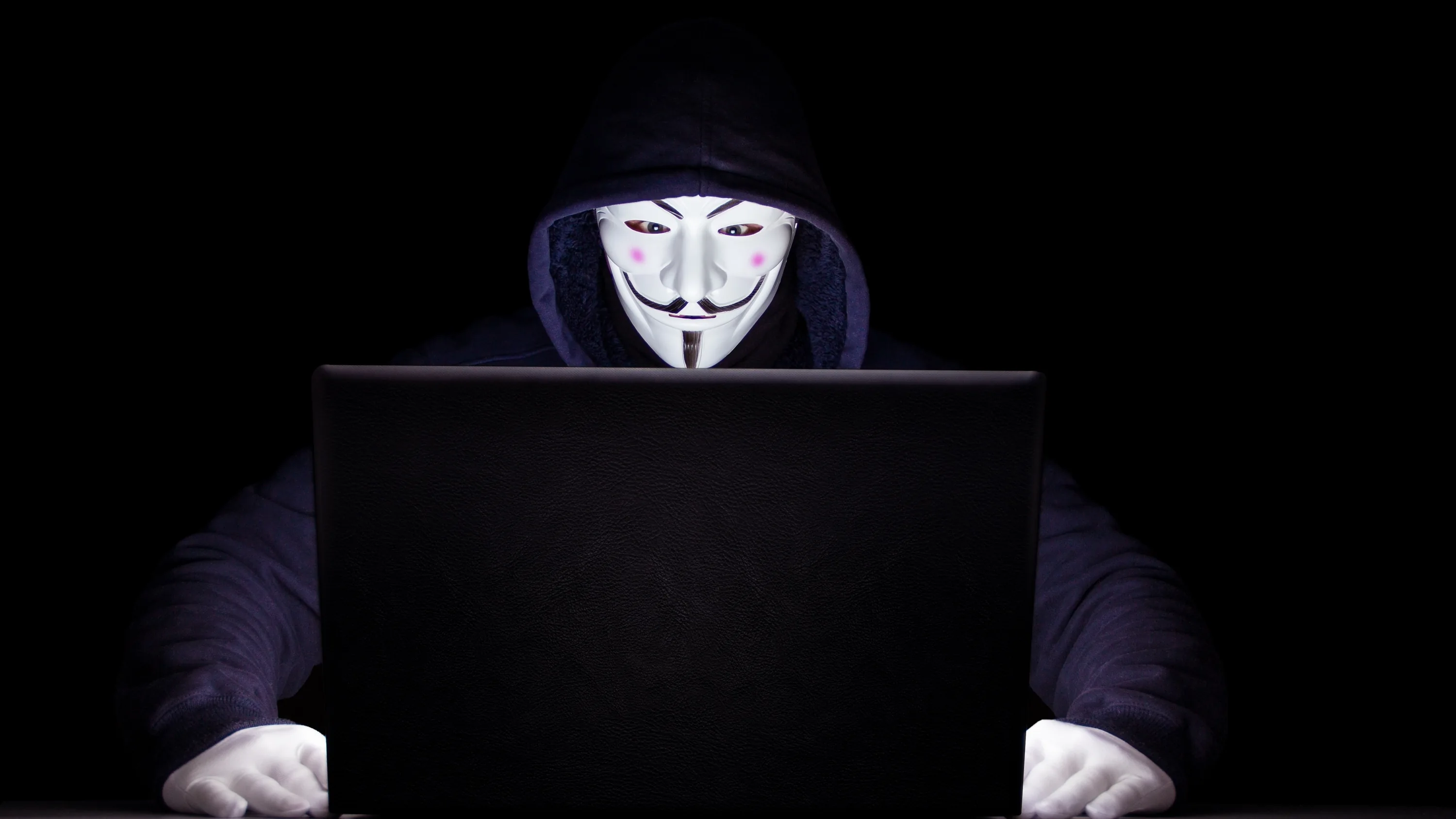 A hooded figure in a pale grinning mask sits behind an open laptop, hands on the desk and the room around them fully black.