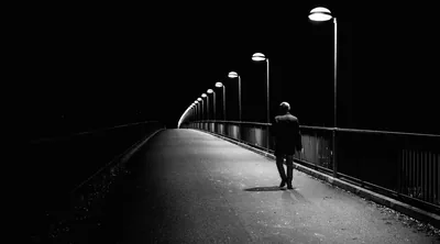 A curved row of streetlamps recedes into darkness as a man in a jacket walks the empty bridge, the left half of the sky staying fully black.