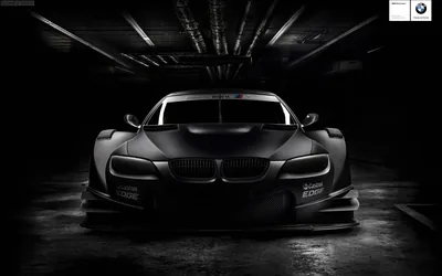 Wide-arch BMW race car facing the camera in a tunnel, strip lights streaking away overhead and monochrome grading pushing everything but the kidney grilles into grey.