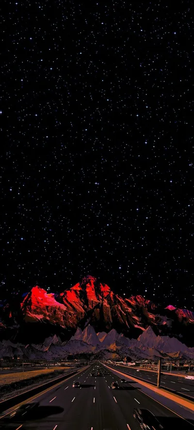 A rocky ridge burns deep red under a thick star field while a lit highway runs straight out of the bottom of the frame.