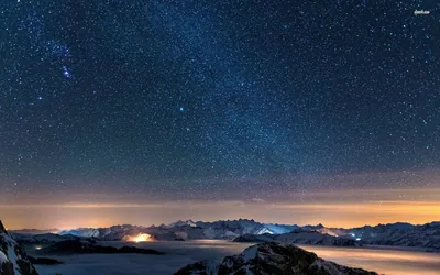 A dense field of stars fills the frame above a snowy mountain chain, orange town glow seeping through valley fog along the horizon.