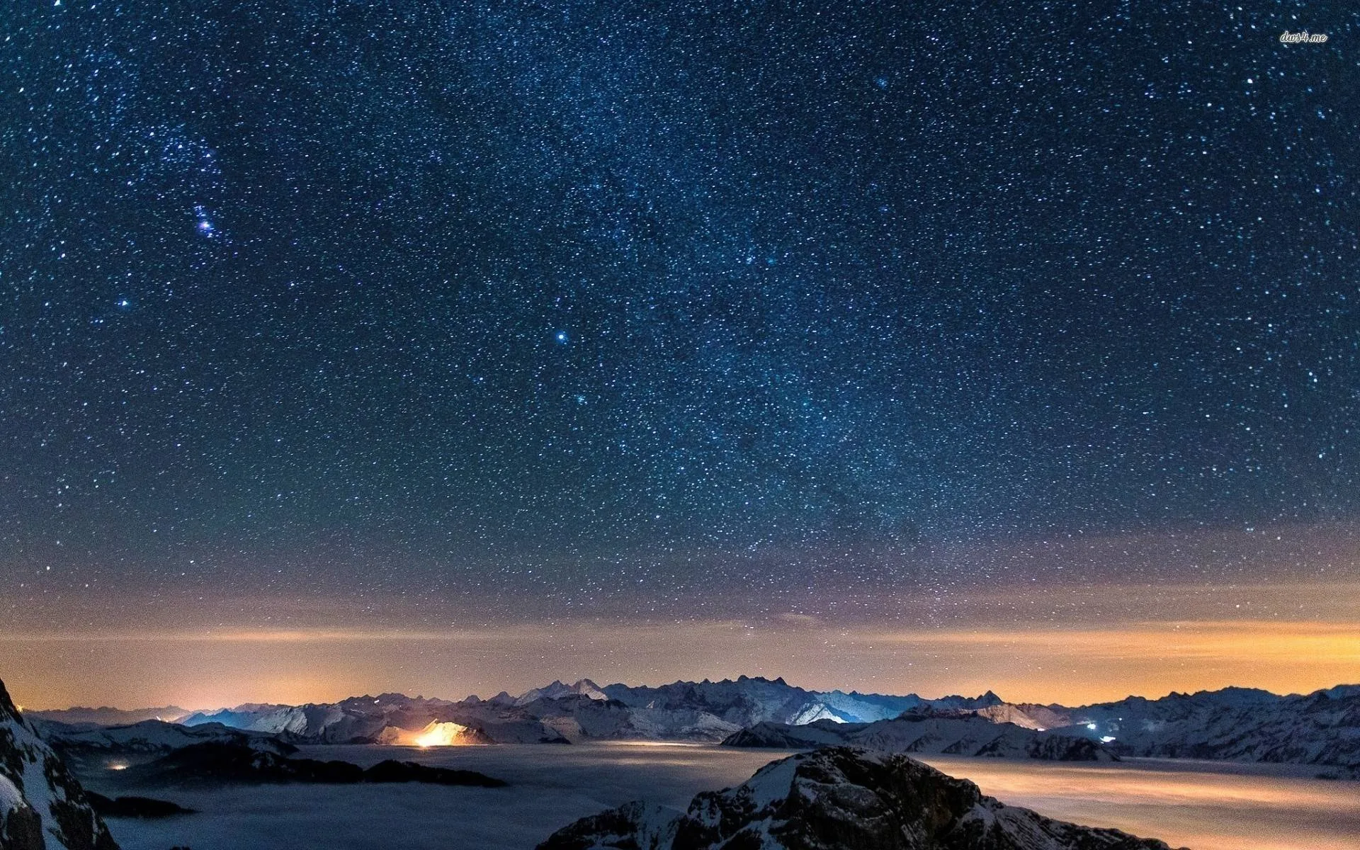 A dense field of stars fills the frame above a snowy mountain chain, orange town glow seeping through valley fog along the horizon.