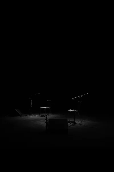 A bare stage with a stool and a low platform sits under one weak spill of light, the rest of the hall swallowed by black.
