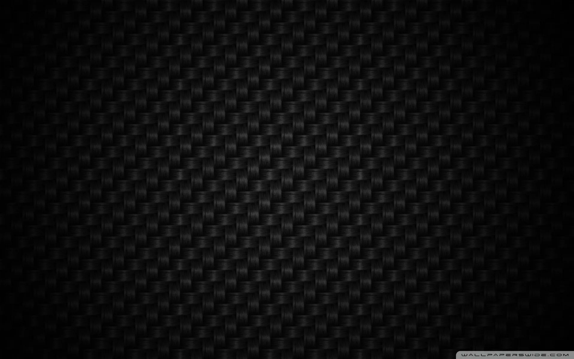 The tight over-under weave of carbon fibre fills the frame, lit from the upper left so the sheen falls off into black at the corners.
