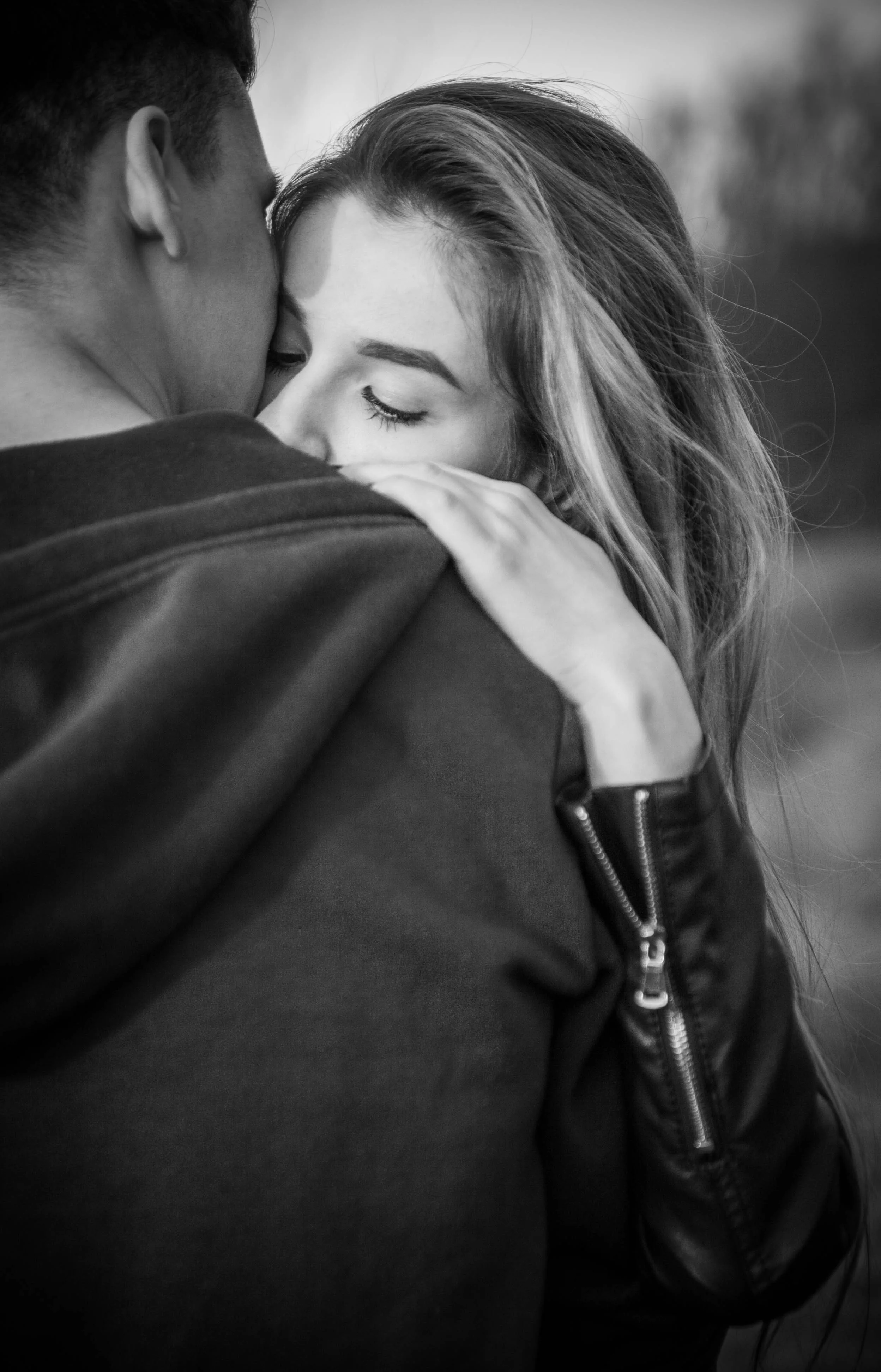 A woman rests her face against a man's shoulder with her eyes closed, her hair falling loose, the black and white frame cropped tight enough to lose the background.