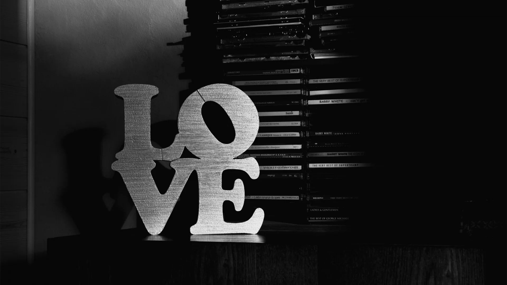 Stacked block letters spelling LOVE sit on a table in a dim room, shot in monochrome with heavy shadow behind them.