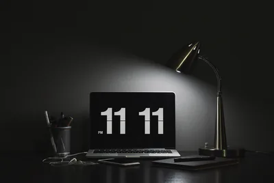 A desk lamp lights a laptop showing the time in large white digits, the room behind it dropping into black.