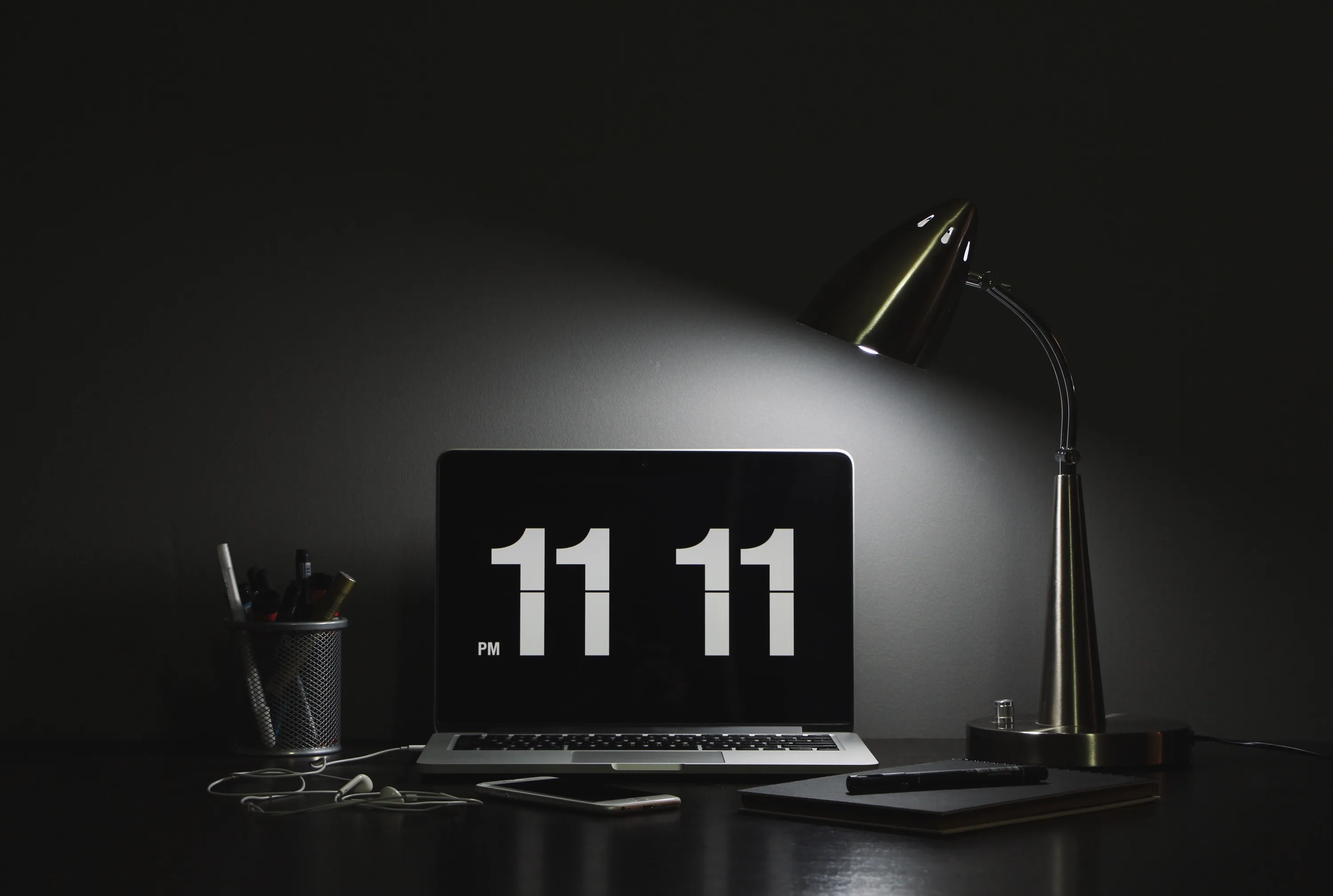 A desk lamp lights a laptop showing the time in large white digits, the room behind it dropping into black.