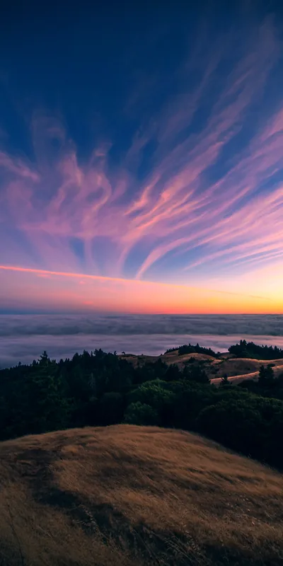 Pink cirrus streaks fan across a twilight sky above a fog-filled valley; the smooth upper half keeps a lock-screen clock readable on this tall frame.