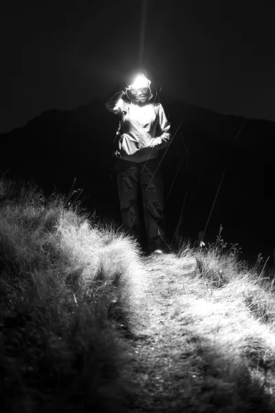 Black and white frame of a walker descending a grassy ridge path, the headlamp burning a bright halo that lights only the trail's bottom third.