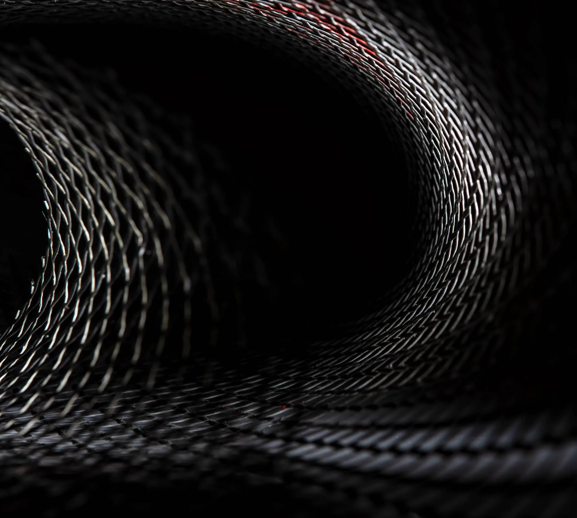 A knitted black mesh curls into a funnel that opens toward the left, one thread of red catching the light where the tunnel narrows.