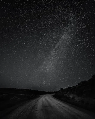 A gravel road runs out of the bottom corner into black hills, the Milky Way climbing diagonally overhead in grainy monochrome.