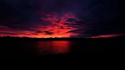 A band of crimson cloud burns above a black treeline and repeats in the flat water below.