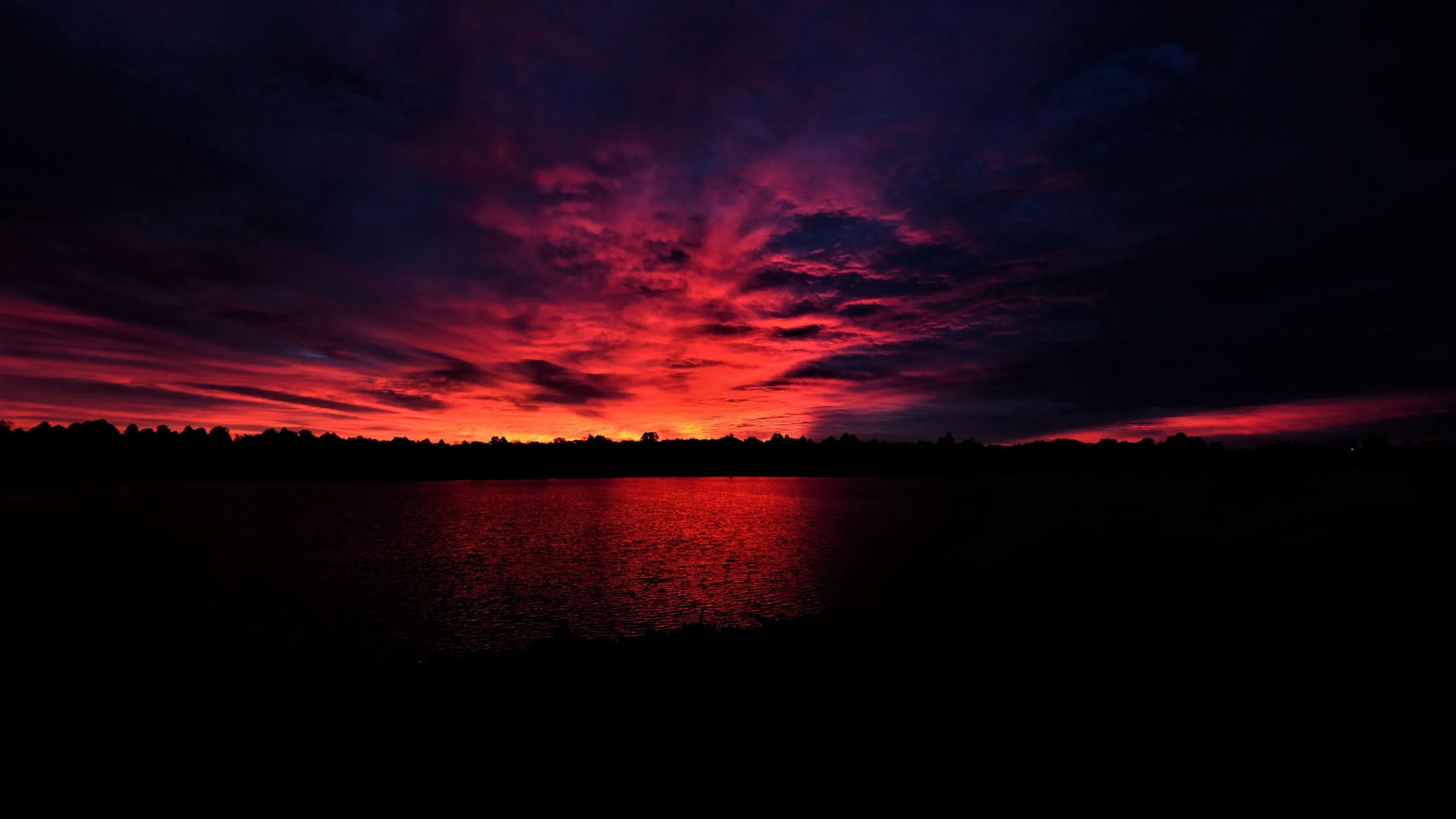 A band of crimson cloud burns above a black treeline and repeats in the flat water below.