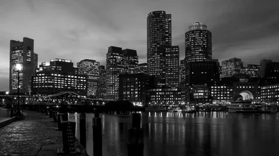 Lit office towers line the far bank in monochrome, old wooden pilings running out along the near edge and window light dropping in soft columns onto the water.
