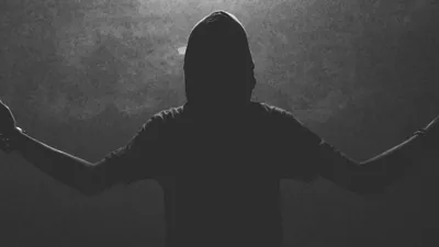A hooded figure stands with both arms flung wide in front of a grainy concrete wall, shot in greyscale with the light coming from behind.
