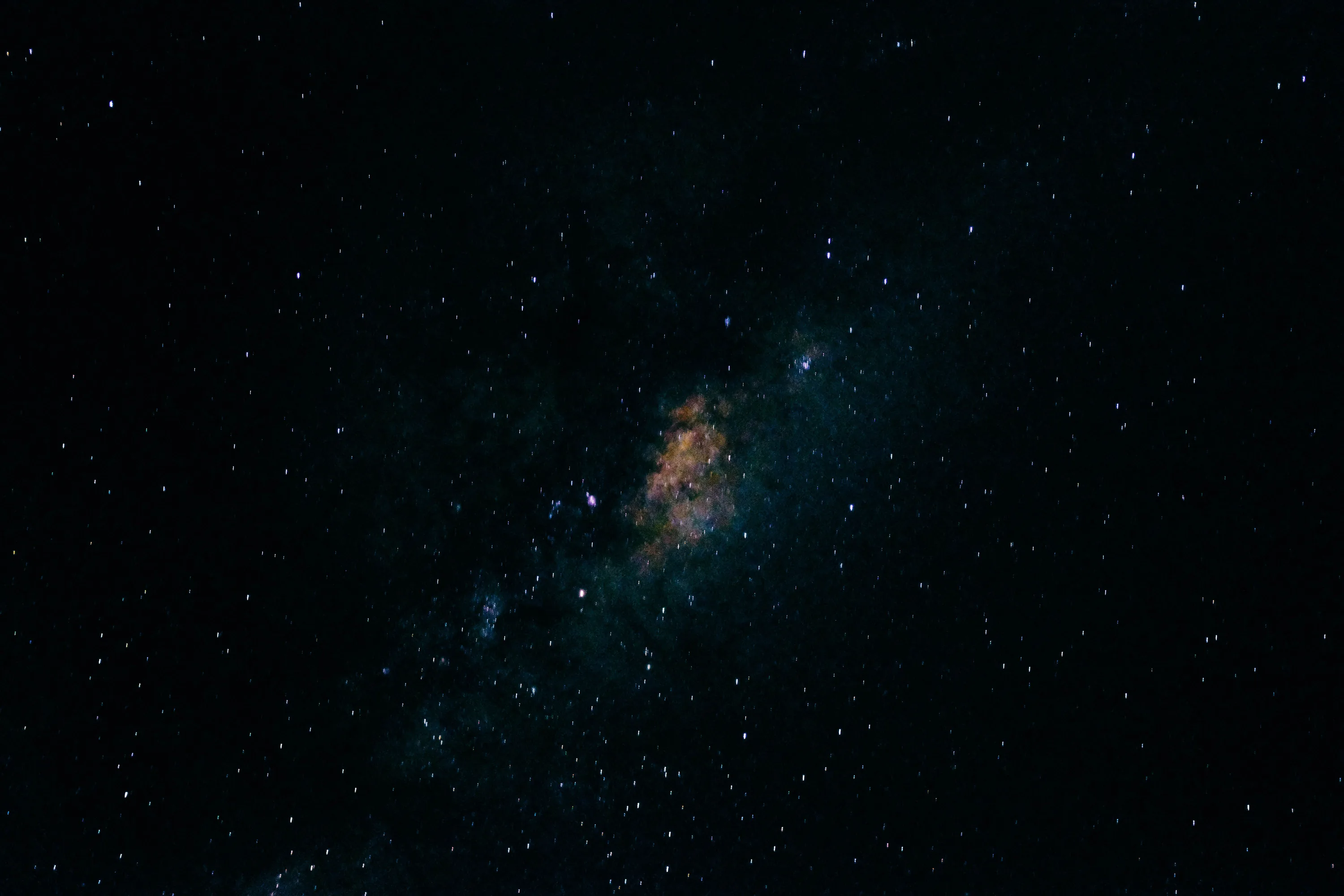 A very dark sky with thousands of small stars and one rust-coloured nebula glowing weakly near the centre.