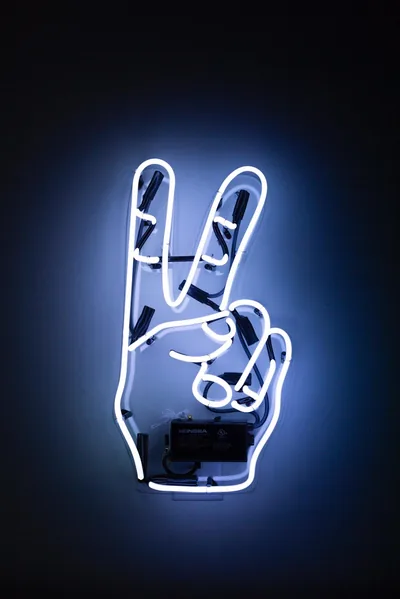 A hand throwing a V sign glows in white and blue tube light, mounted on a dark wall that catches a faint halo behind it.
