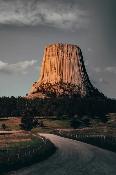 Evening sun catches the columned face of Devils Tower while the road below curves in from the left; grey sky fills the whole upper third.
