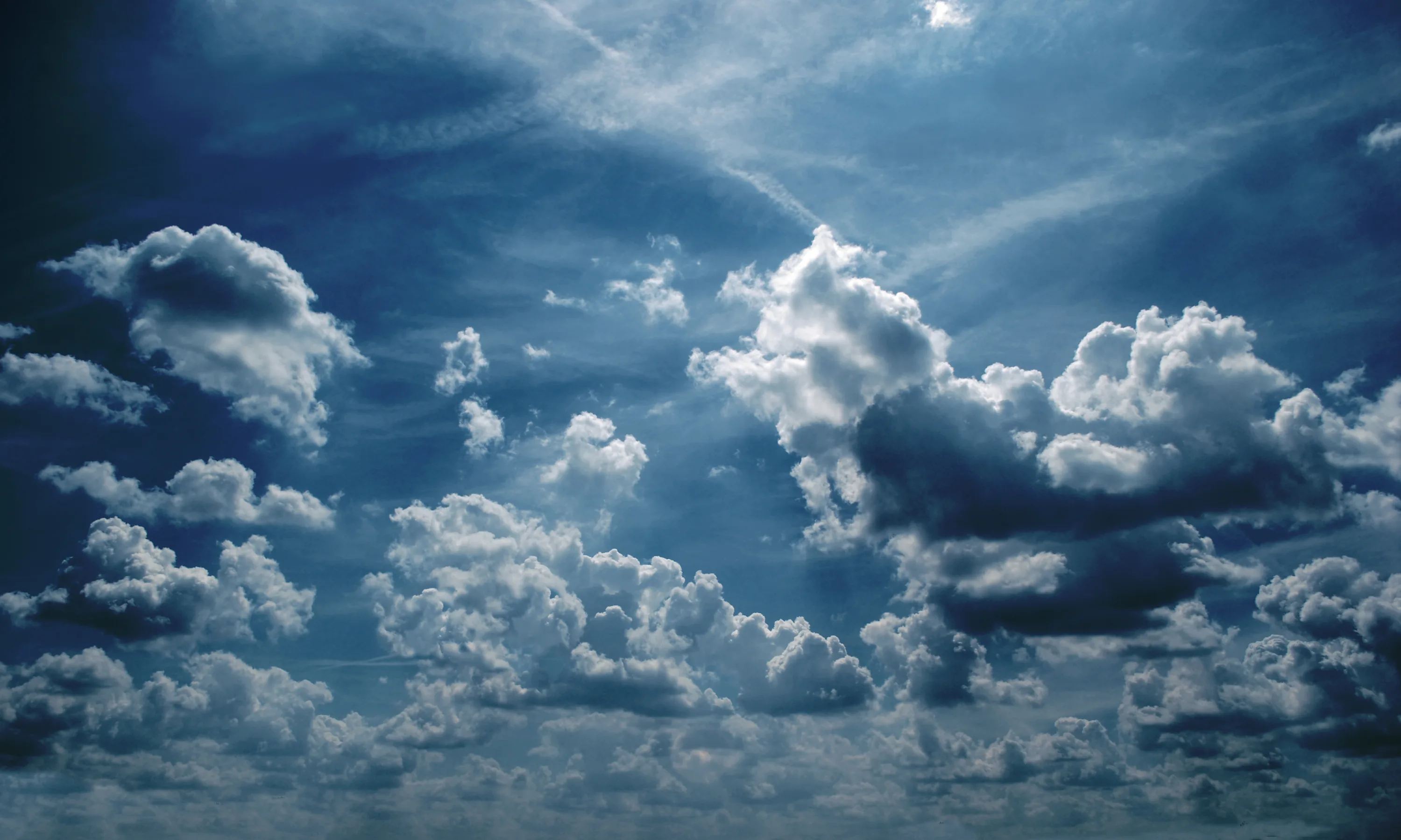 Small moody cumulus scatter across a steel-blue sky, two faint contrails crossing near the top — the whole frame reads almost monochrome.