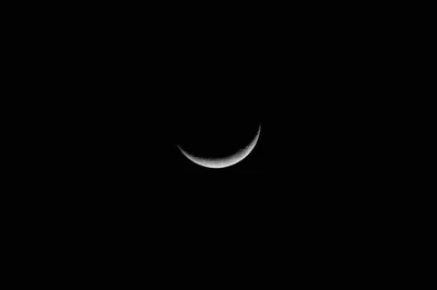 A narrow crescent hangs slightly left of centre in a completely black sky, bright along the rim with the dark limb just readable beside it.