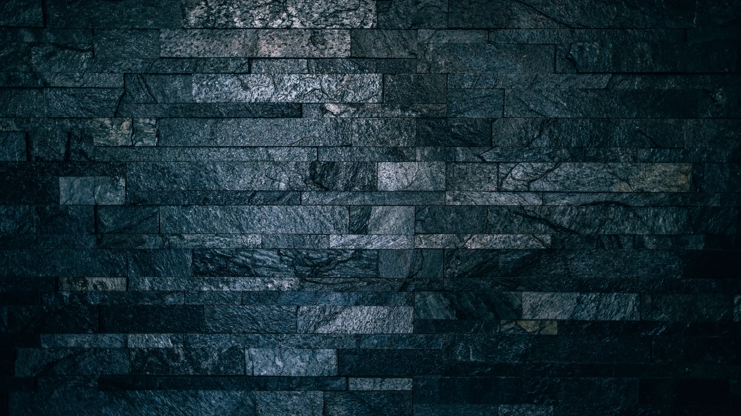 Rough slate strips stack across the wall in cold blue-grey, the light pooling near the centre and the corners sunk in shadow.