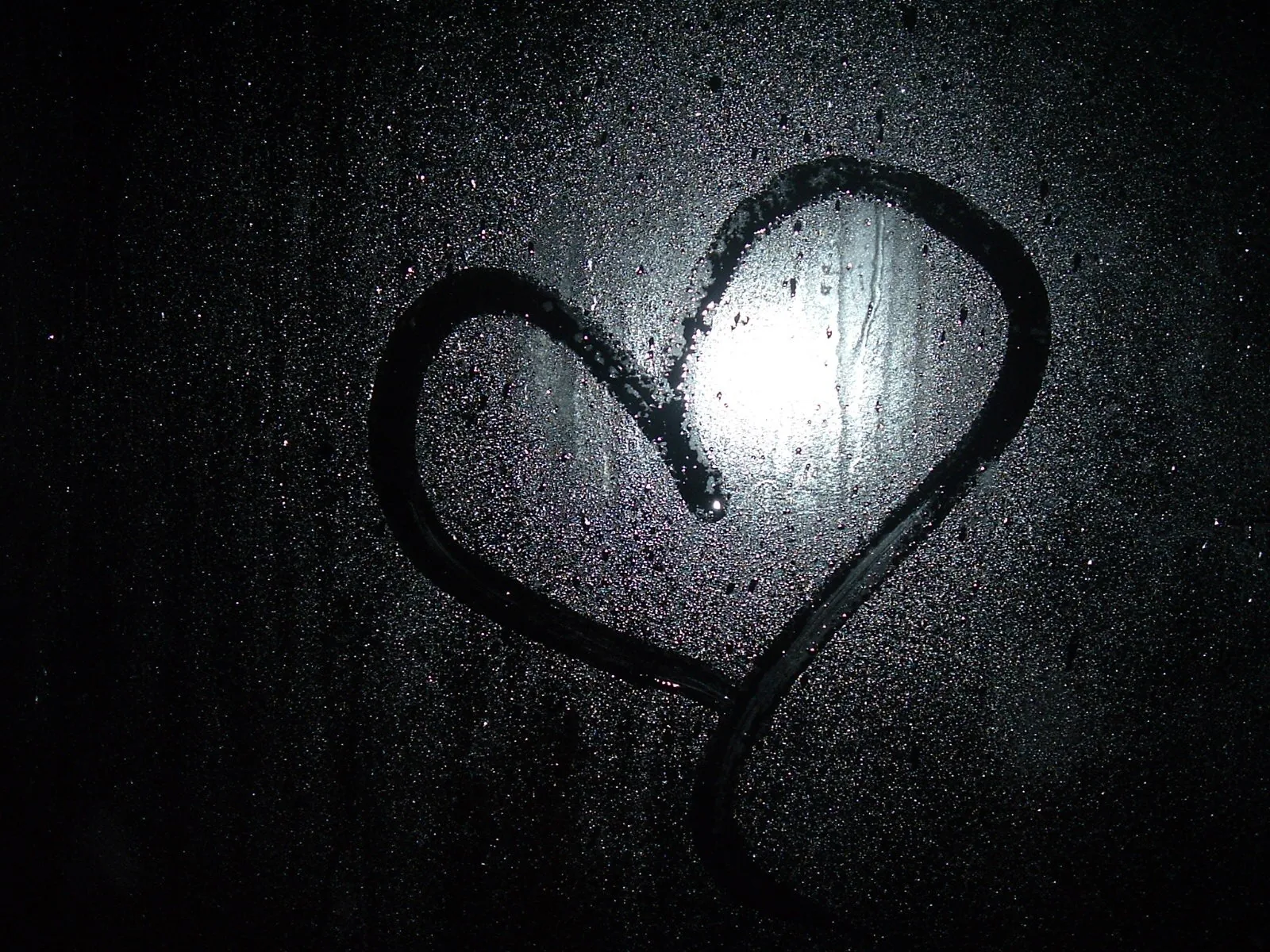 A finger-drawn heart on a misted window at night, one streetlight blurring through the condensation just inside the shape.