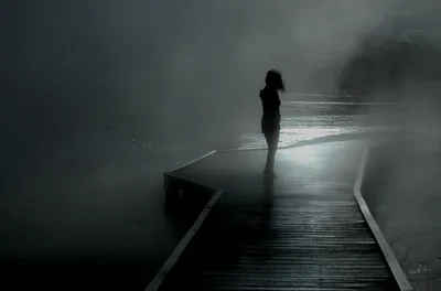 A woman stands mid-boardwalk as fog swallows the planks behind her, the single bright patch of light glowing low on the left of the frame.