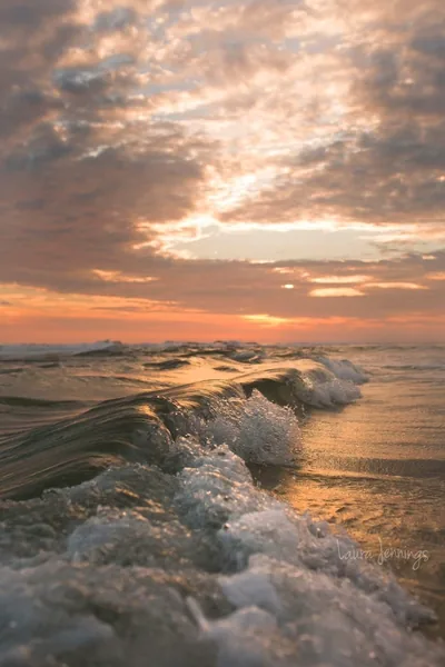 Shot from the waterline as foam tumbles toward the lens, peach and grey clouds glowing overhead — the soft top third keeps a status bar readable.
