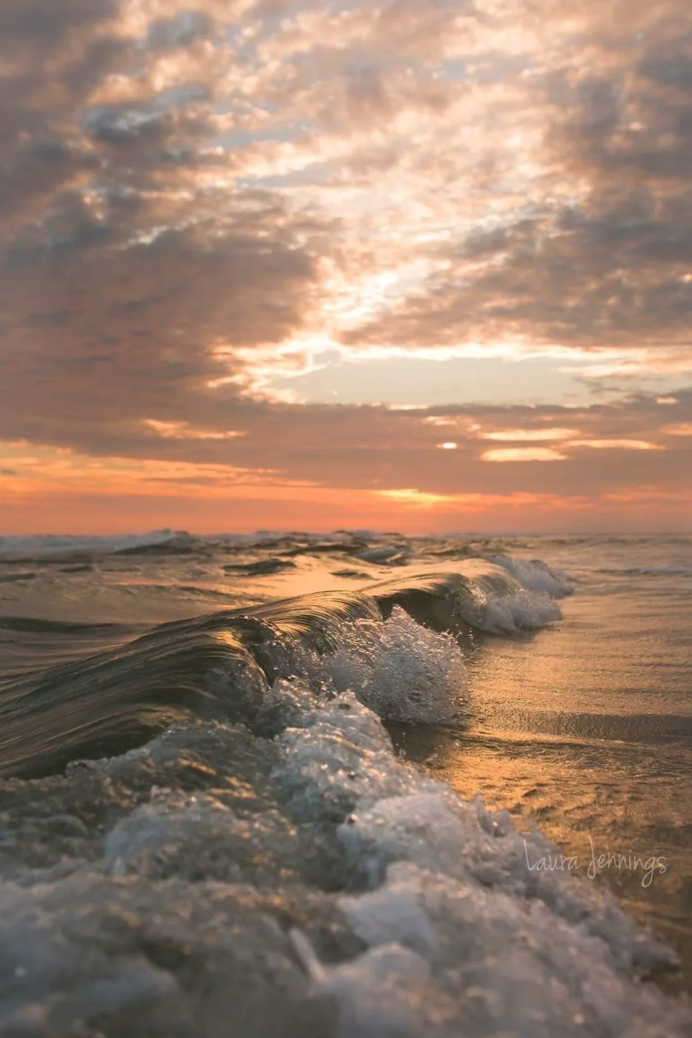Shot from the waterline as foam tumbles toward the lens, peach and grey clouds glowing overhead — the soft top third keeps a status bar readable.