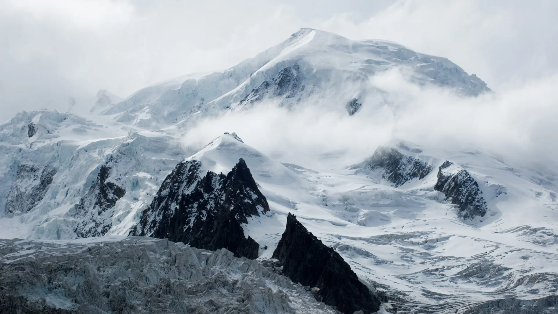 Wind drags cloud across a glaciated summit above black rock spires, the whole scene held in cold near-monochrome greys and whites.