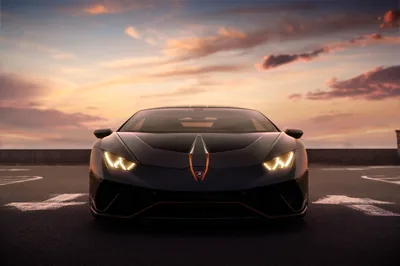 The Lamborghini sits square-on across a rooftop car park, hooked headlights lit and orange trim along the splitter picking up the last colour in the clouds.